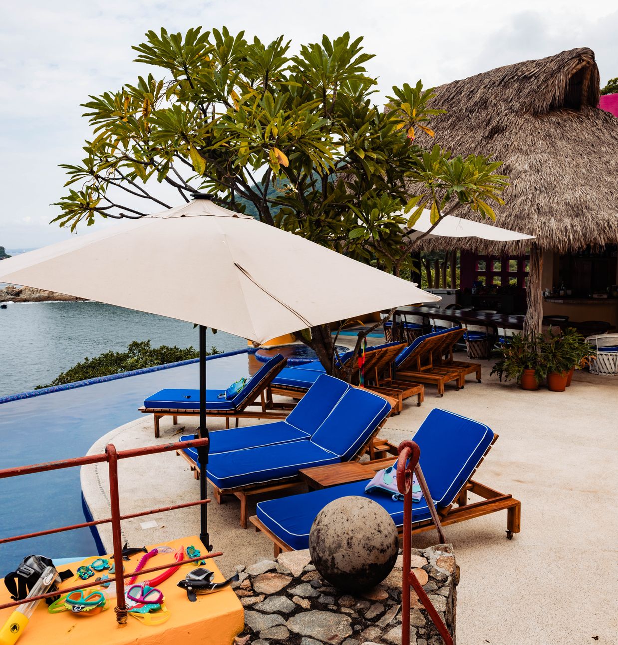 Blue loungers and a white umbrella on a terrace facing the ocean and islets, with a palapa roof alongside