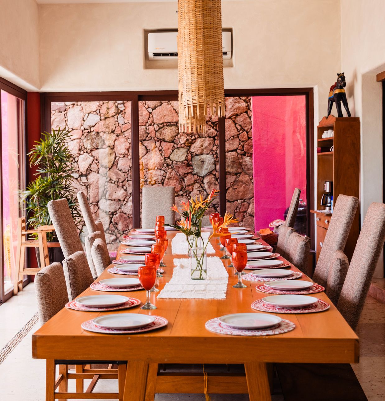 Long dining table set for a large group with red glasses, plates and flowers in a room with pink and stone walls