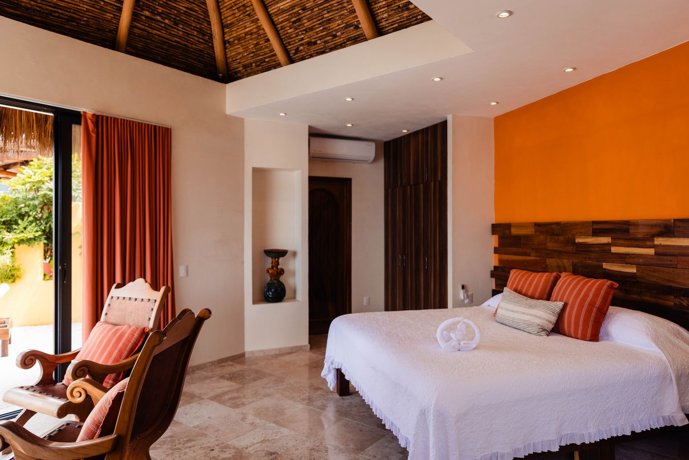 Bedroom with an orange accent wall, wood headboard, palapa ceiling and a rocking chair at Villa Mandarinas