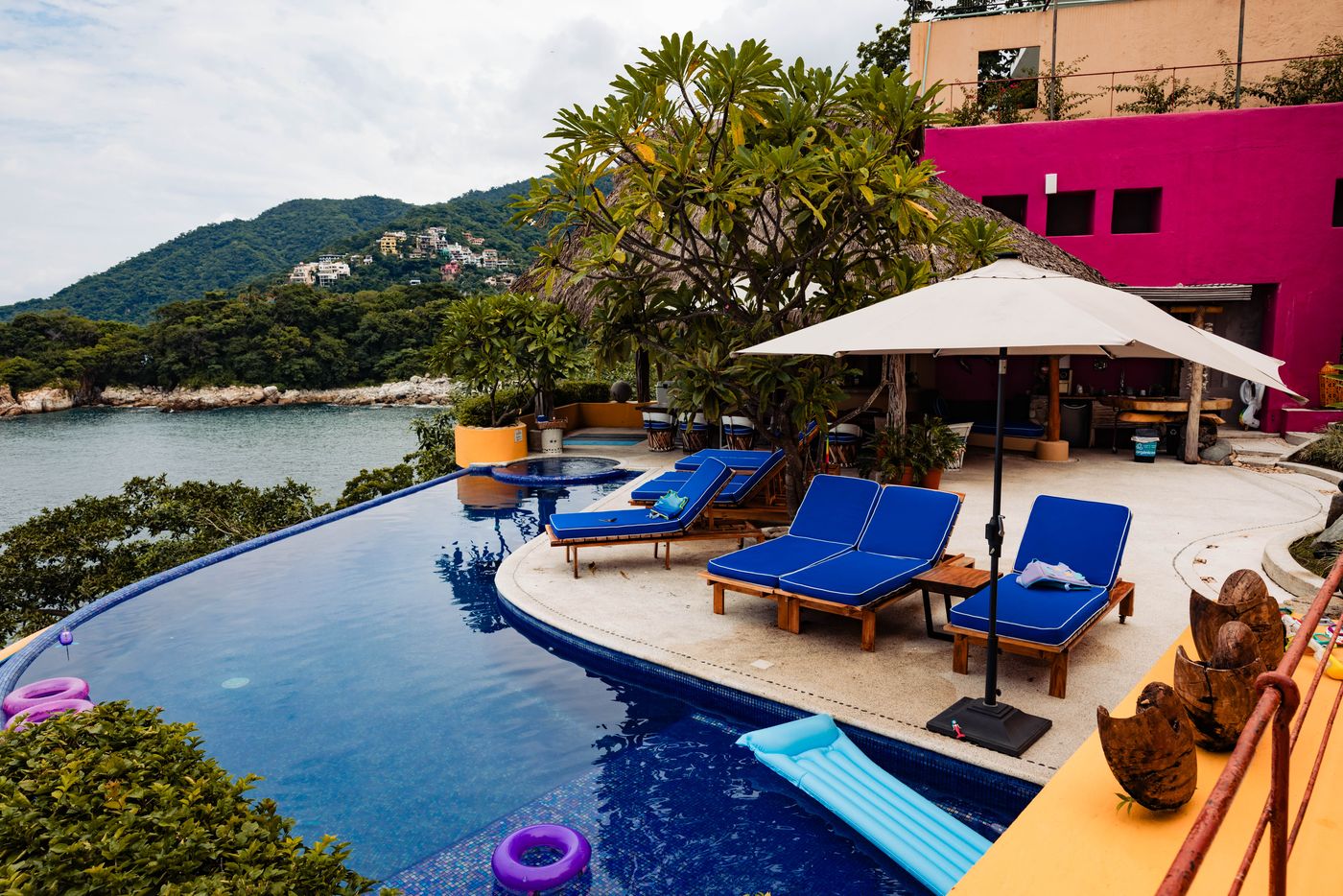 Infinity pool deck with blue loungers and an umbrella overlooking the bay, with a pink building behind